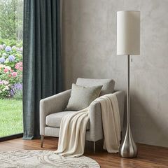 68" silver floor lamp with a white fabric drum shade beside a gray armchair and a cozy blanket in a bright room.