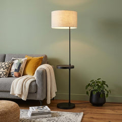 64" beige and black tray table floor lamp with a beige fabric drum shade, placed beside a cozy sofa and a potted plant.