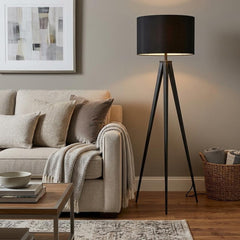 Black Metal Tripod Floor Lamp with Black Fabric Drum Shade, illuminating a cozy living room with a beige sofa and decorative 