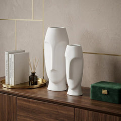 Set of two ceramic white cylinder table vases with abstract face designs, displayed on a wooden surface with decor items.