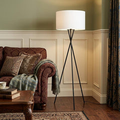 66" Black Tripod Floor Lamp with White Fabric Drum Shade, positioned next to a cozy sofa in a warmly decorated living room.