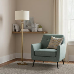 63" Gold Column Floor Lamp with Off White Metal Drum Shade beside a teal armchair on a textured rug in a cozy room setting.