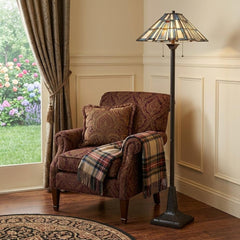 62" Bronze Two Light Traditional Floor Lamp with Gray and Ivory Abstract Tiffany Glass Shade beside a cozy armchair and throw