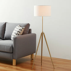 Gold Metal Adjustable Tripod Floor Lamp with White Linen Drum Shade beside a gray sofa, featuring a decorative pillow and woo