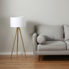 Gold metal adjustable tripod floor lamp with white linen drum shade beside a light gray sofa with a round cushion.