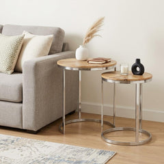 Set of two natural and silver solid wood and iron round frame nested tables beside a gray sofa, featuring decorative items.